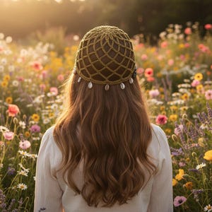 May include: An olive green crocheted cap featuring a net-like pattern and small white cowrie shells dangling from the bottom edge. The cap is worn on long, wavy brown hair, with the back of the head and shoulders visible in the frame.
