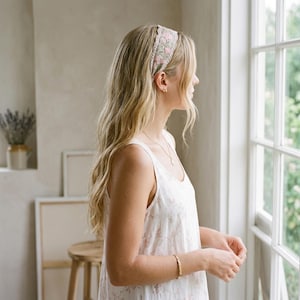 May include: A fabric headband featuring a light-colored base with an embroidered pattern of pink flowers and green leaves. The headband has a scalloped edge design and is worn across the top of the head, resting against long blonde hair.