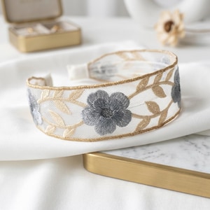 May include: A wide headband made of sheer white mesh fabric featuring embroidered grey flowers and gold metallic leaf vines. The edges of the headband are finished with a gold metallic trim, creating a structured and decorative hair accessory.