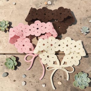 May include: Three crocheted flower chokers in shades of brown, pink, and cream. The chokers are arranged on a wooden surface with small succulents and pebbles. Each choker features a floral design and a tie closure.