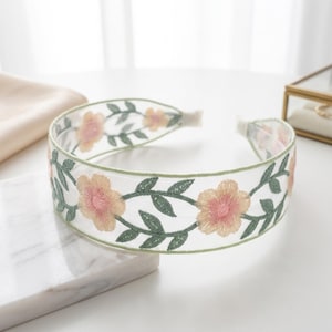 May include: A wide headband made of sheer white mesh fabric featuring an embroidered pattern of light pink flowers with dark pink centers and green leaves. The edges of the headband are finished with a thin green trim along the top and bottom.