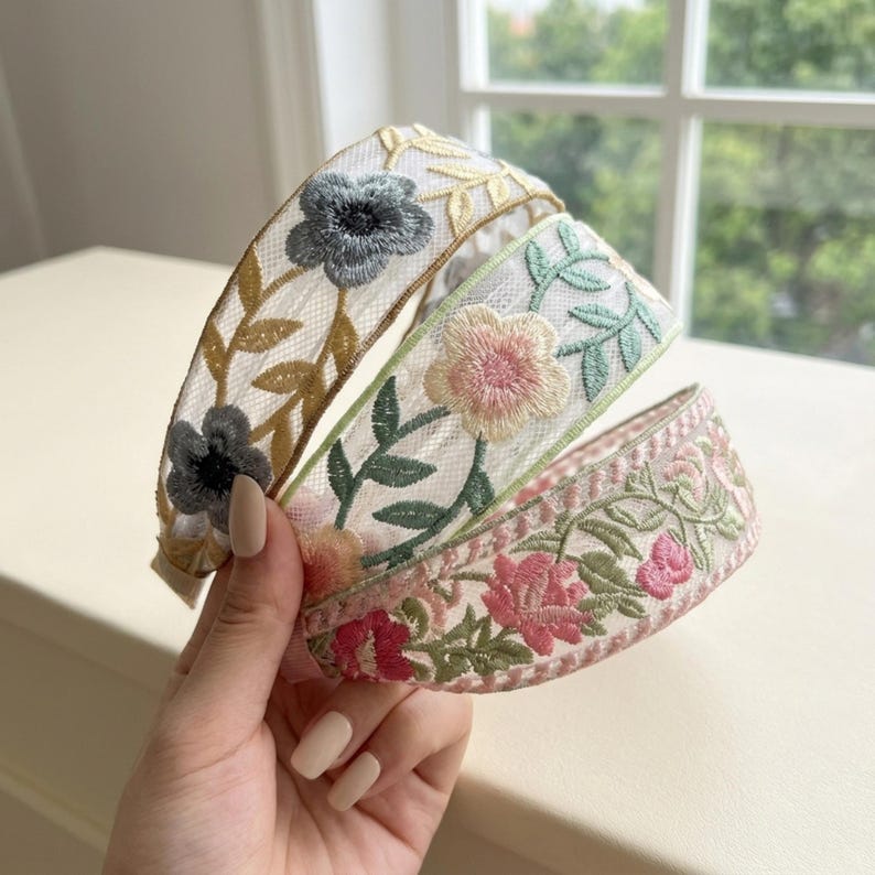 May include: Three headbands made of sheer white mesh fabric featuring intricate floral embroidery. The first headband displays grey flowers with gold vines, the second shows pink flowers with green leaves, and the third features pink and red floral patterns.