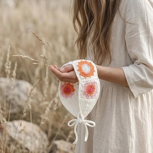 May include: A white crochet headband featuring three colorful flower designs in orange, pink, and light blue. The headband is tied with a white ribbon. The background is a field of tall, dry grass.