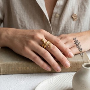 May include: A gold-toned snake ring featuring a textured, scale-like pattern on the body and a smooth, tapered tail. The ring wraps around the finger in a spiral design. A small gold-toned pin shaped like a raised fist with a female symbol is attached to the beige shirt.