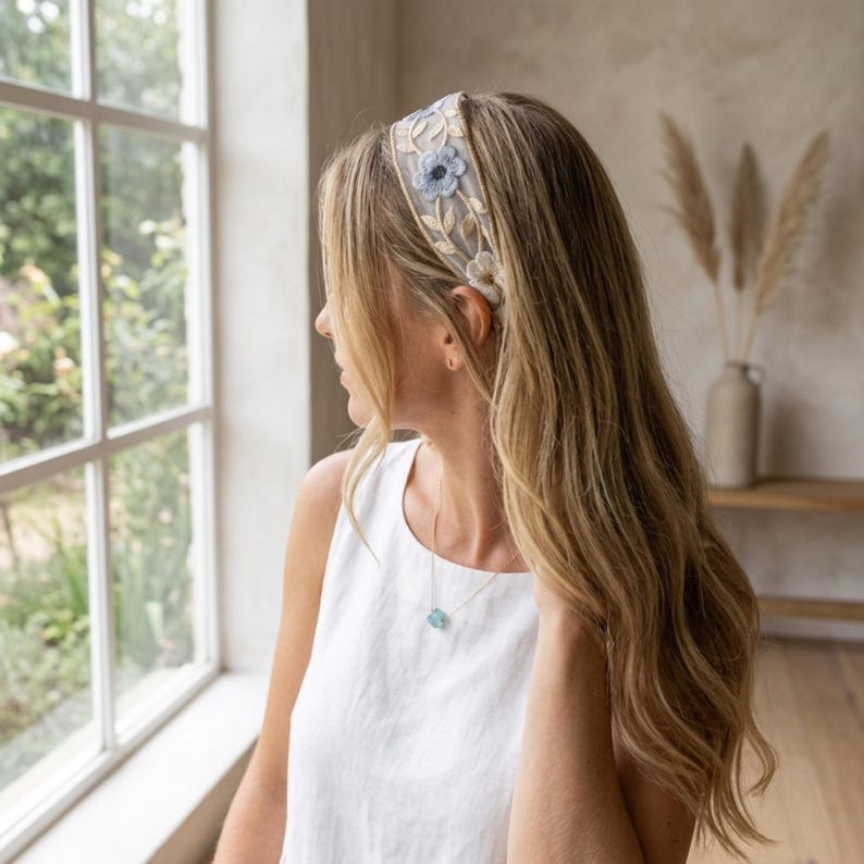 May include: A sheer headband featuring delicate embroidery of light blue and white flowers with green stems on a translucent mesh base. The floral pattern is evenly spaced across the band, which is worn on long, wavy, light brown hair.