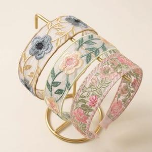 May include: Three headbands made of sheer mesh fabric featuring intricate floral embroidery. The first headband displays blue flowers and gold vines, the second shows peach flowers with green leaves, and the third features pink flowers and green foliage.