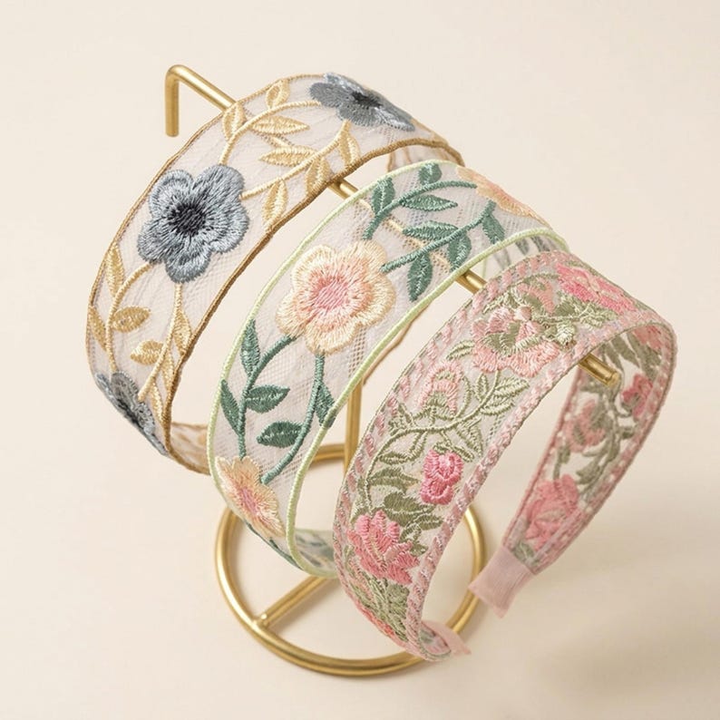 May include: Three headbands made of sheer mesh fabric featuring intricate floral embroidery. The first headband displays blue flowers and gold vines, the second shows peach flowers with green leaves, and the third features pink flowers and green foliage.
