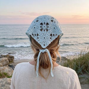 May include: A light blue crochet headscarf featuring a detailed floral motif pattern. The triangular head covering is tied at the nape of the neck with two thin, matching crochet strings, creating a secure fit over the back of the head.