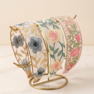 May include: Three headbands displayed on a gold metal stand. The first features blue flowers and gold leaves on white mesh. The second has pink flowers and green vines on white mesh. The third has pink floral embroidery on a white fabric band.