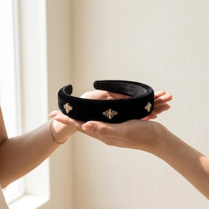 May include: A wide, black velvet headband featuring evenly spaced, small gold-toned bee embellishments. The headband has a thick, padded structure and a smooth, soft-textured surface, with the metallic bee motifs attached along the outer band.