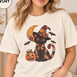 Könnte beinhalten: Ein elfenbeinfarbenes Kurzarm-T-Shirt mit einer Grafik einer schwarzen Katze, die einen Patchwork-Hexenhut und eine Fliege trägt. Die Katze sitzt neben einem Kürbis, im Hintergrund sind eine Mondsichel und kleine Fledermäuse zu sehen. Text IVORY.