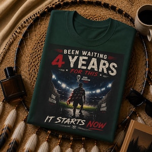 May include: Dark green t-shirt with the text "Been Waiting 4 Years For This." The graphic shows a person with a flag on a soccer field. The text "It Starts Now" is at the bottom. The shirt has a crew neck.