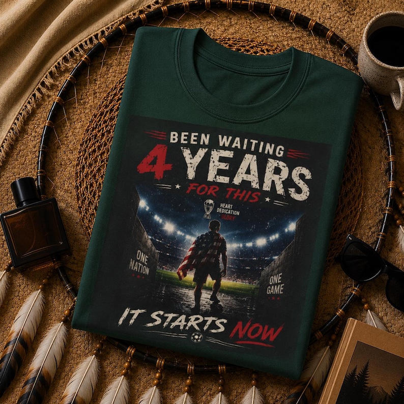 May include: Dark green t-shirt with the text "Been Waiting 4 Years For This." The graphic shows a person with a flag on a soccer field. The text "It Starts Now" is at the bottom. The shirt has a crew neck.