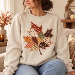 Puede incluir: Una sudadera de cuello redondo en color beige con un gran gráfico central de una hoja de arce compuesta por varios patrones de tela tipo patchwork en tonos naranja, marrón, verde y crema, incluyendo motivos florales, de cuadros y de calabazas.