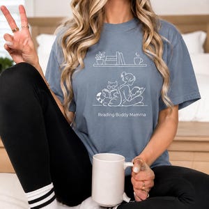 Può includere: T-shirt blu polveroso con grafica bianca di una persona che legge con un gatto, libri e una tazza di caffè. Il testo "Reading Buddy Mamma" è sotto la grafica. La persona indossa leggings neri e calzini bianchi con strisce nere.