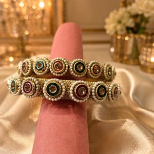 May include: Two gold-toned bangles featuring a textured surface, adorned with a repeating pattern of circular accents in shades of red, green, and blue, each centered with a clear stone and bordered by a ring of small white faux pearls.