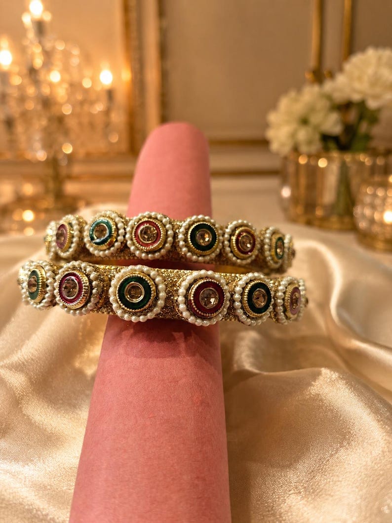 May include: Two gold-toned bangles featuring a textured surface, adorned with a repeating pattern of circular accents in shades of red, green, and blue, each centered with a clear stone and bordered by a ring of small white faux pearls.