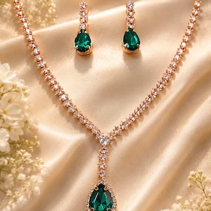 Pode incluir: Um conjunto de joias composto por um colar e um par de brincos pendentes. O colar apresenta uma corrente dourada com cristais transparentes e uma pedra central verde-esmeralda em forma de gota. Os brincos possuem pedras verdes a condizer.