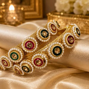 May include: Two gold-toned bangles featuring a textured surface and a series of circular motifs. Each motif contains a central clear stone surrounded by a ring of red or green enamel, bordered by a delicate row of small white faux pearls.