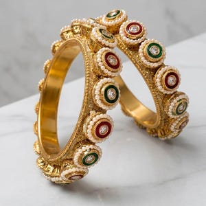 May include: Two gold-toned bangles featuring a textured surface and a series of circular accents. Each accent contains a central clear stone surrounded by a ring of small white beads, alternating between deep red and dark green enamel borders.