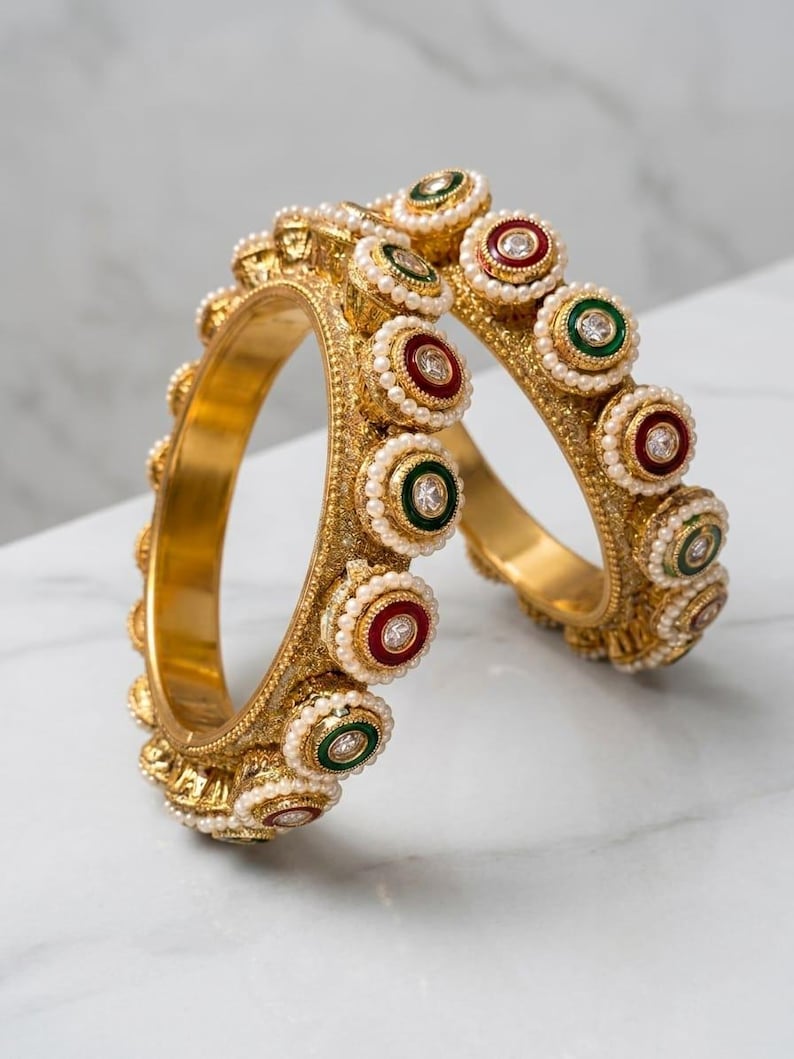 May include: Two gold-toned bangles featuring a textured surface and a series of circular accents. Each accent contains a central clear stone surrounded by a ring of small white beads, alternating between deep red and dark green enamel borders.