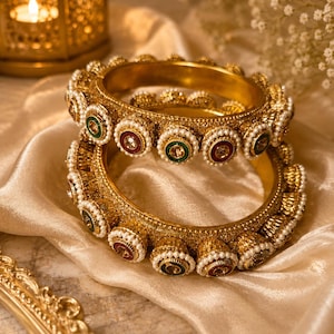 May include: Two gold-toned metal bangles featuring a textured surface, adorned with a repeating pattern of small white faux pearls surrounding circular insets in alternating deep red and dark green colors with a central clear stone.
