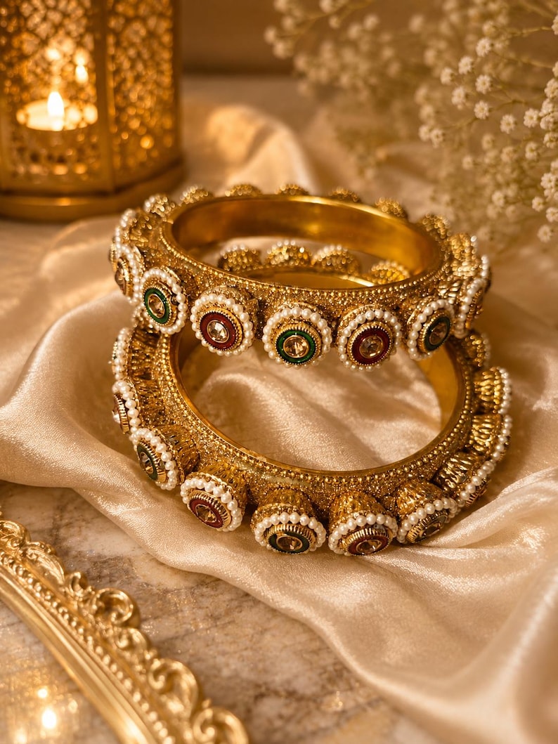 May include: Two gold-toned metal bangles featuring a textured surface, adorned with a repeating pattern of small white faux pearls surrounding circular insets in alternating deep red and dark green colors with a central clear stone.