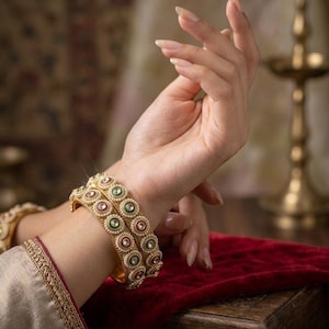 May include: Two gold-toned bangles featuring a repeating circular pattern with alternating red and green centers, surrounded by small white beads. The bangles have a textured metallic finish and are worn on the wrist, showcasing a detailed design.