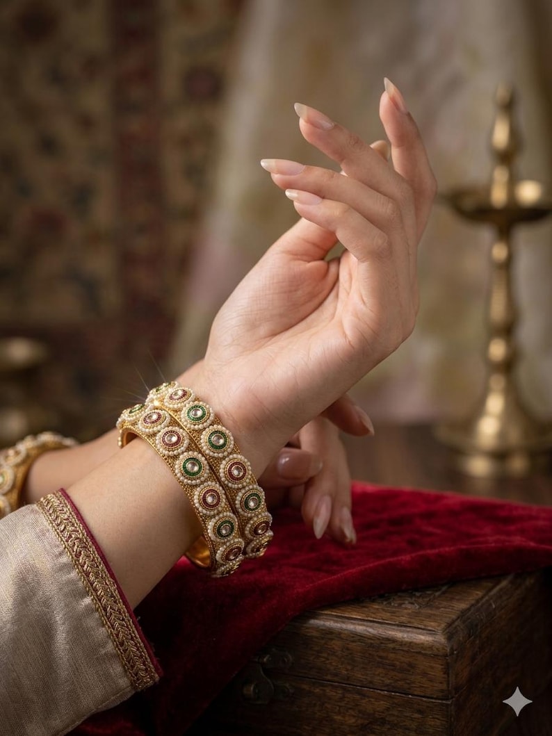 May include: Two gold-toned bangles featuring a repeating circular pattern with alternating red and green centers, surrounded by small white beads. The bangles have a textured metallic finish and are worn on the wrist, showcasing a detailed design.