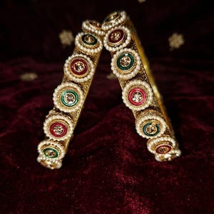 May include: Two gold-toned bangles featuring a series of circular ornaments. Each ornament is bordered by small white faux pearls and contains a central stone in either deep red or dark green, set against a metallic gold textured band.