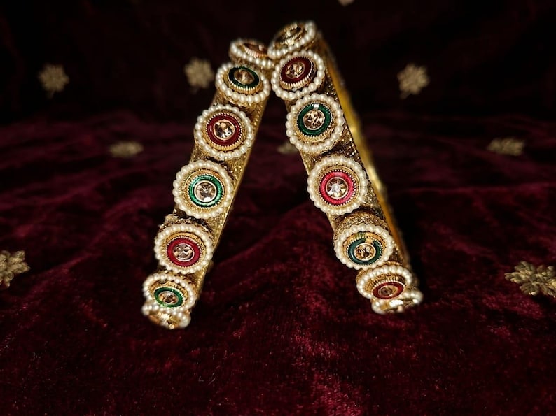May include: Two gold-toned bangles featuring a series of circular ornaments. Each ornament is bordered by small white faux pearls and contains a central stone in either deep red or dark green, set against a metallic gold textured band.