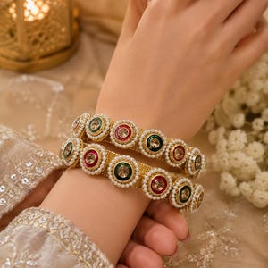 May include: Two gold-toned bangles featuring a textured surface and a repeating pattern of circular motifs. Each motif contains a central clear stone surrounded by alternating red or green enamel borders and a delicate frame of small white pearls.