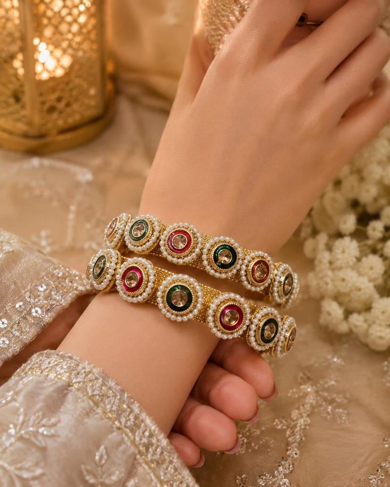 May include: Two gold-toned bangles featuring a textured surface and a repeating pattern of circular motifs. Each motif contains a central clear stone surrounded by alternating red or green enamel borders and a delicate frame of small white pearls.