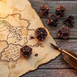 May include: A collection of bronze-colored polyhedral dice with floral designs, arranged on a vintage map. A quill pen with a feather is positioned near the map. The dice are numbered, and the map has a detailed, aged appearance.