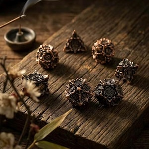 May include: A set of bronze-colored polyhedral dice arranged on a weathered wooden surface. The dice have intricate floral designs and are used for tabletop games. An incense burner and sprig of flowers are in the background.