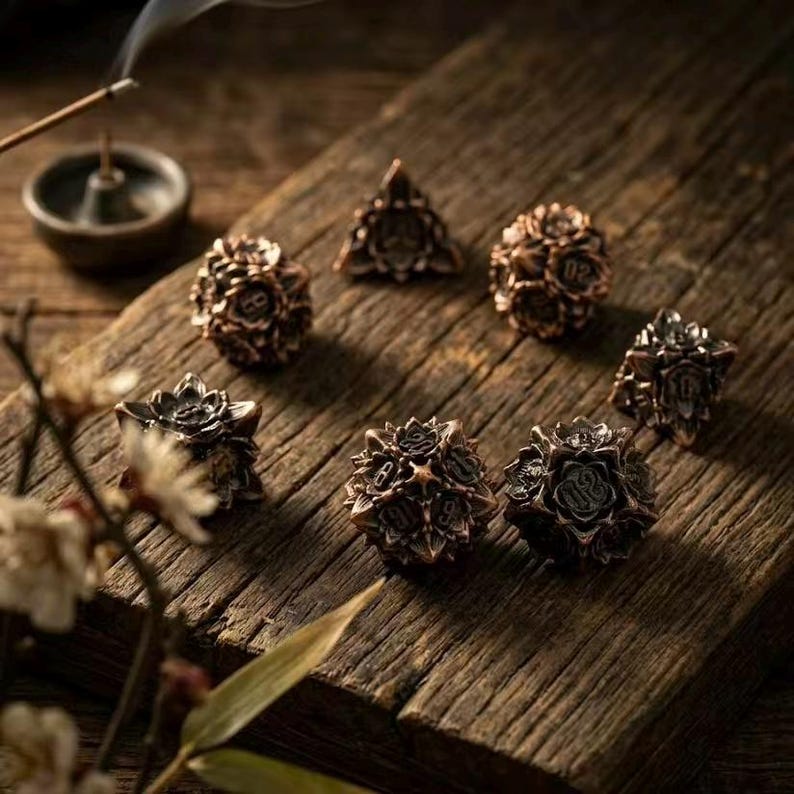 May include: A set of bronze-colored polyhedral dice arranged on a weathered wooden surface. The dice have intricate floral designs and are used for tabletop games. An incense burner and sprig of flowers are in the background.