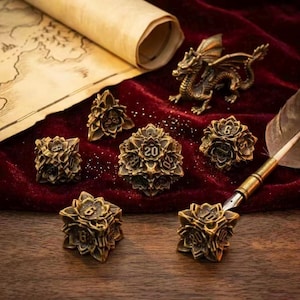 May include: A collection of antique gold-colored polyhedral dice with a floral design, perfect for tabletop games. The set includes a D20, D12, D10, D8, D6, and D4. A small bronze dragon figurine and a scroll are in the background.