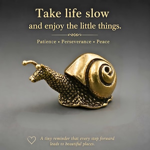 Peut inclure: Une figurine d'escargot en métal doré avec un corps texturé et une coquille lisse en spirale. Le texte au-dessus indique : Take life slow and enjoy the little things. Patience • Perseverance • Peace. En dessous : A tiny reminder that every step forward leads to beautiful places.