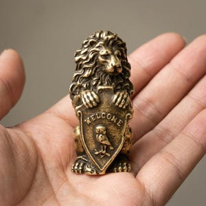 Peut inclure: Une petite figurine de lion en métal couleur laiton, représentée debout. Le lion tient un bouclier portant le mot "WELCOME" en lettres en relief au-dessus d'une gravure de hibou. La surface présente une finition texturée de style ancien.