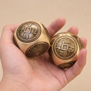 Puede incluir: Dos dados de metal color latón con inserciones circulares en cada cara. Las inserciones muestran caracteres chinos en relieve y patrones tradicionales similares a monedas con un acabado oscuro y antiguo. Superficie dorada pulida.