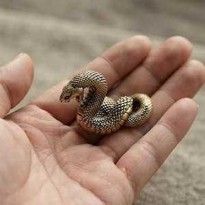 以下が含まれることがあります： 真鍮色の仕上げが施された金属素材で作られた、小さなとぐろを巻いたヘビの置物です。表面全体に詳細な隆起した鱗の質感が施されており、頭部はわずかに持ち上がり、前方を向いた姿勢で湾曲しています。