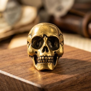 Puede incluir: Una pequeña figura de calavera de metal en color dorado con cuencas oculares, cavidad nasal y una hilera de dientes detallados. La calavera tiene una superficie pulida y reflectante, moldeada con rasgos anatómicos realistas y pómulos definidos.