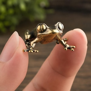 Könnte beinhalten: Eine kleine Froschfigur aus Metall mit einem brünierten Gold- und Dunkelbraun-Finish. Der Frosch ist in einer kauernden Position dargestellt, mit markanten, abgerundeten Augen, detaillierten Schwimmhäuten und einem glatten, gewölbten Rücken.