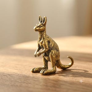 Peut inclure: Une petite figurine de kangourou debout en laiton avec une finition dorée antique patinée. La sculpture présente des textures détaillées sur le pelage, des oreilles pointues, une queue courbée et une poche, représentant l'animal debout.