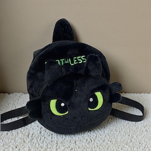 Könnte beinhalten: Schwarzer Plüschrucksack in Form eines Drachenkopfes mit großen grünen Augen und dem Wort "Toothless" in Grün. Inklusive schwarzer Schultergurte.