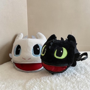 May include: Two plush bags shaped like dragon heads. One is white with blue and black accents, and the other is black with green and black accents. Both have red-lined zippered mouths and small carrying straps.
