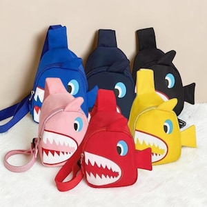 Pode incluir: Uma coleção de bolsas tipo bandoleira em forma de tubarão em várias cores: azul, rosa, marinho, vermelho, amarelo e preto. Cada bolsa apresenta um design de rosto de tubarão com dentes brancos, olhos azuis e uma barbatana. As bolsas têm alças ajustáveis.