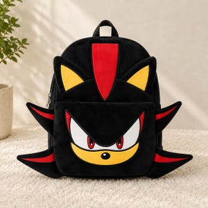 Może przedstawiać: Czarny plecak z pluszowym wzorem postaci Shadow the Hedgehog. Torba posiada żółte uszy, czerwony pasek oraz przednią kieszeń z białymi oczami, czerwonymi źrenicami i żółtym pyszczkiem postaci z czarnymi akcentami.