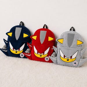 May include: Three plush backpacks in dark blue, red, and light grey. Each bag features a stylised character face with yellow ears, a silver forehead patch, and a circular patch at the bottom with the text QIANLONG ALL STAR and a star icon.