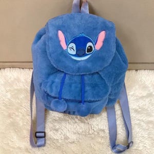 May include: A blue plush backpack featuring the character Stitch from Lilo & Stitch. The backpack has adjustable straps, a top handle, and a flap closure. The character's face is embroidered on the front, with pink ears and a winking eye.